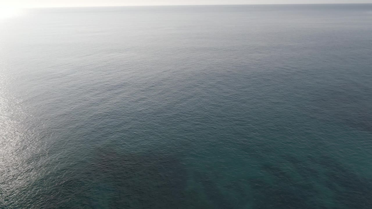 Wide aerial view at Buzzards Beach East Cape BCS Mexico showing open blue ocean, a faint horizon, darker submerged formations, soft glare, and no visible people.
