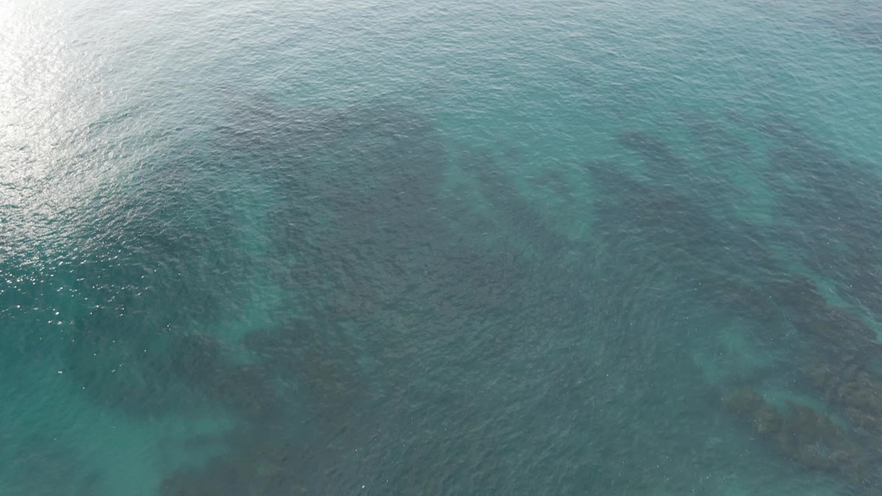 Aerial frame over Buzzards Beach East Cape BCS Mexico with blue-green water, a broad darker underwater reef zone on the left, surface shimmer, and no visible people.