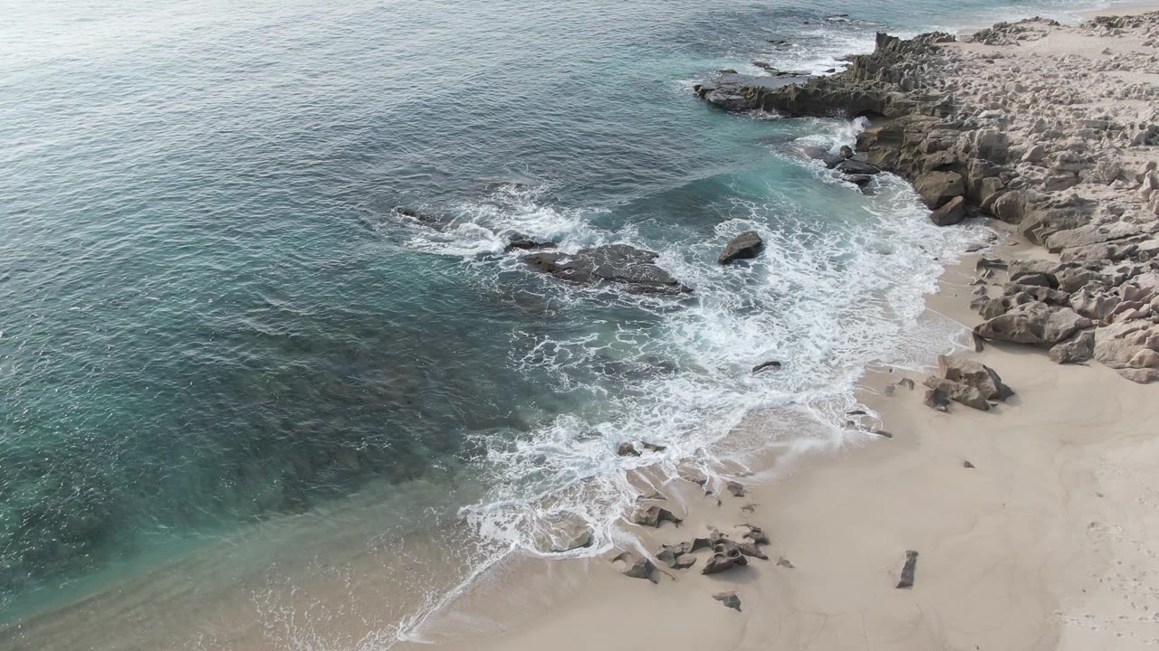 Aerial frame from Buzzards Beach East Cape BCS Mexico showing blue-green water, white surf, dark shoreline rocks, pale sand, and no visible people.