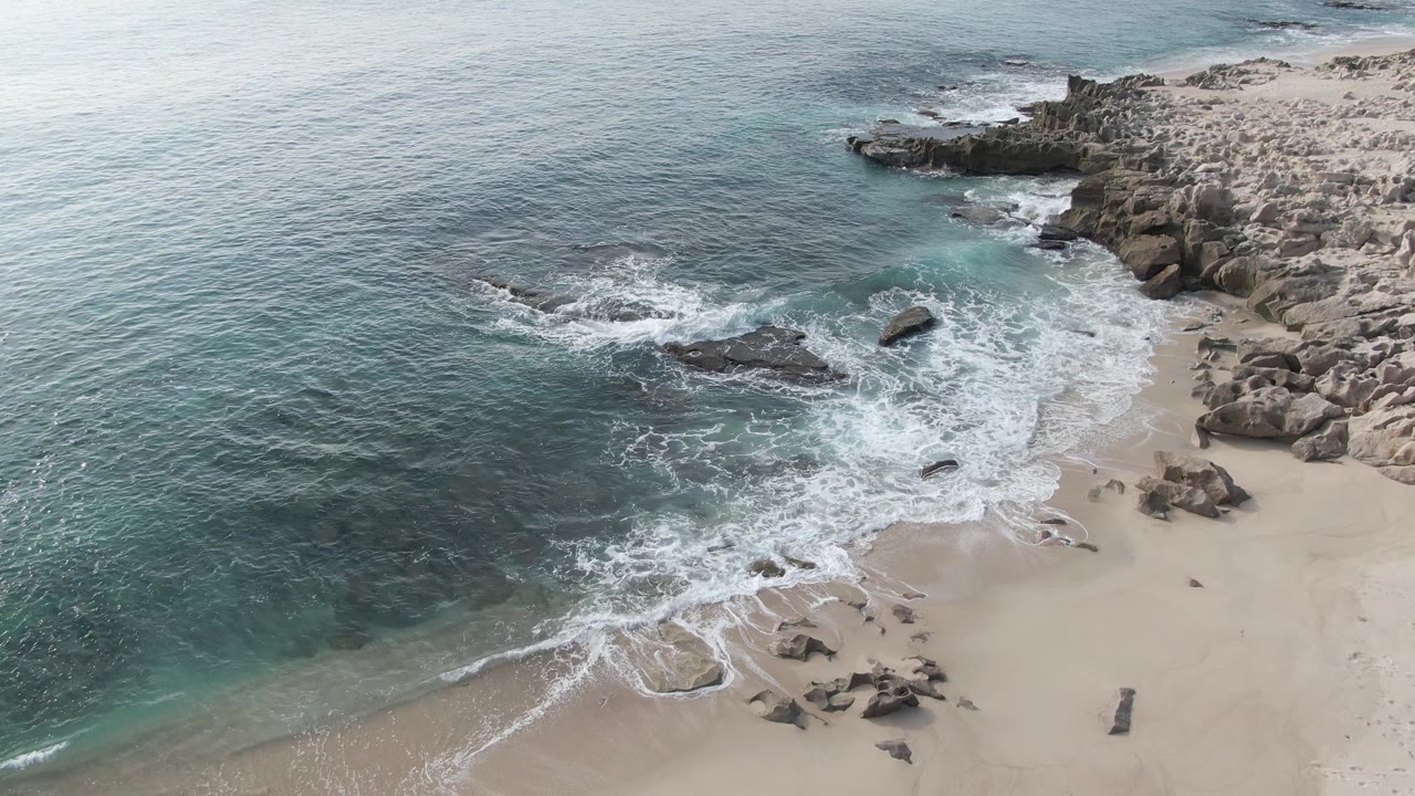 Aerial view at Buzzards Beach East Cape BCS Mexico with surf spreading farther across the sand, open water on the left, rocky outcrops on the right, and no visible people.