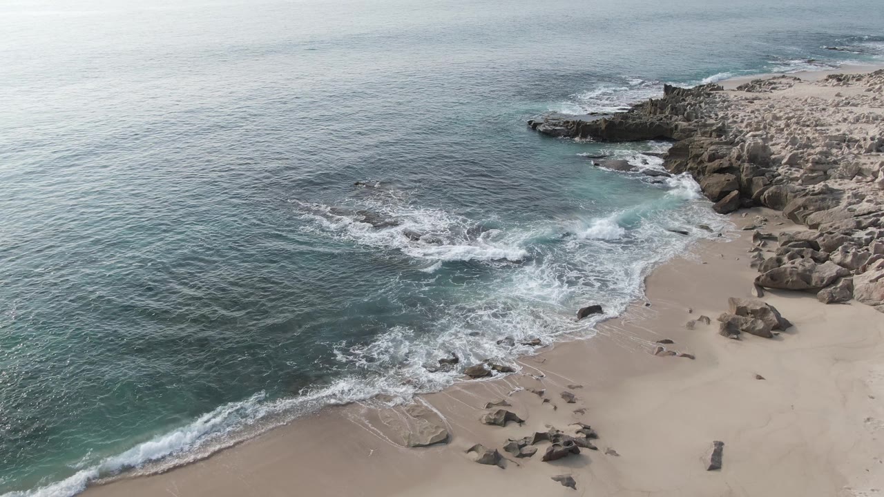 Aerial shoreline view with waves crossing sand and rocks.