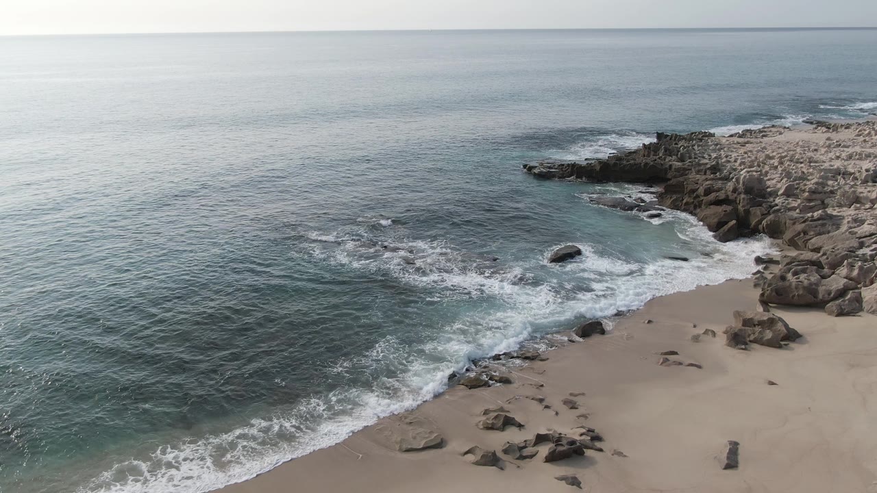 At Buzzards Beach East Cape BCS Mexico, blue-gray ocean water meets a sandy shore with rocks along the right edge and no visible people.