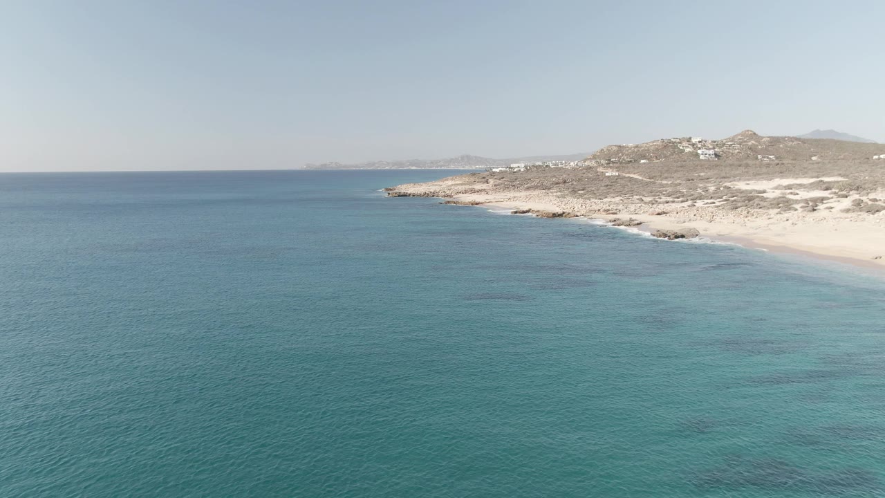 Second frame of Buzzards Beach East Cape BCS Mexico with clear blue-green water covering most of the view, the beach angled along the right edge, rocky terrain beyond it, and no visible people.