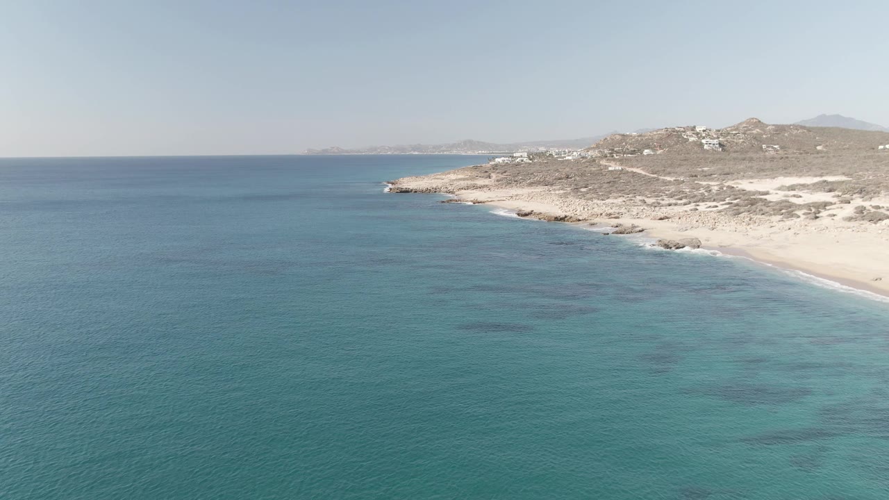 Aerial frame over Buzzards Beach East Cape BCS Mexico showing turquoise water, pale sand, rocky hillside, scattered white buildings, and no visible people at the start of the clip.