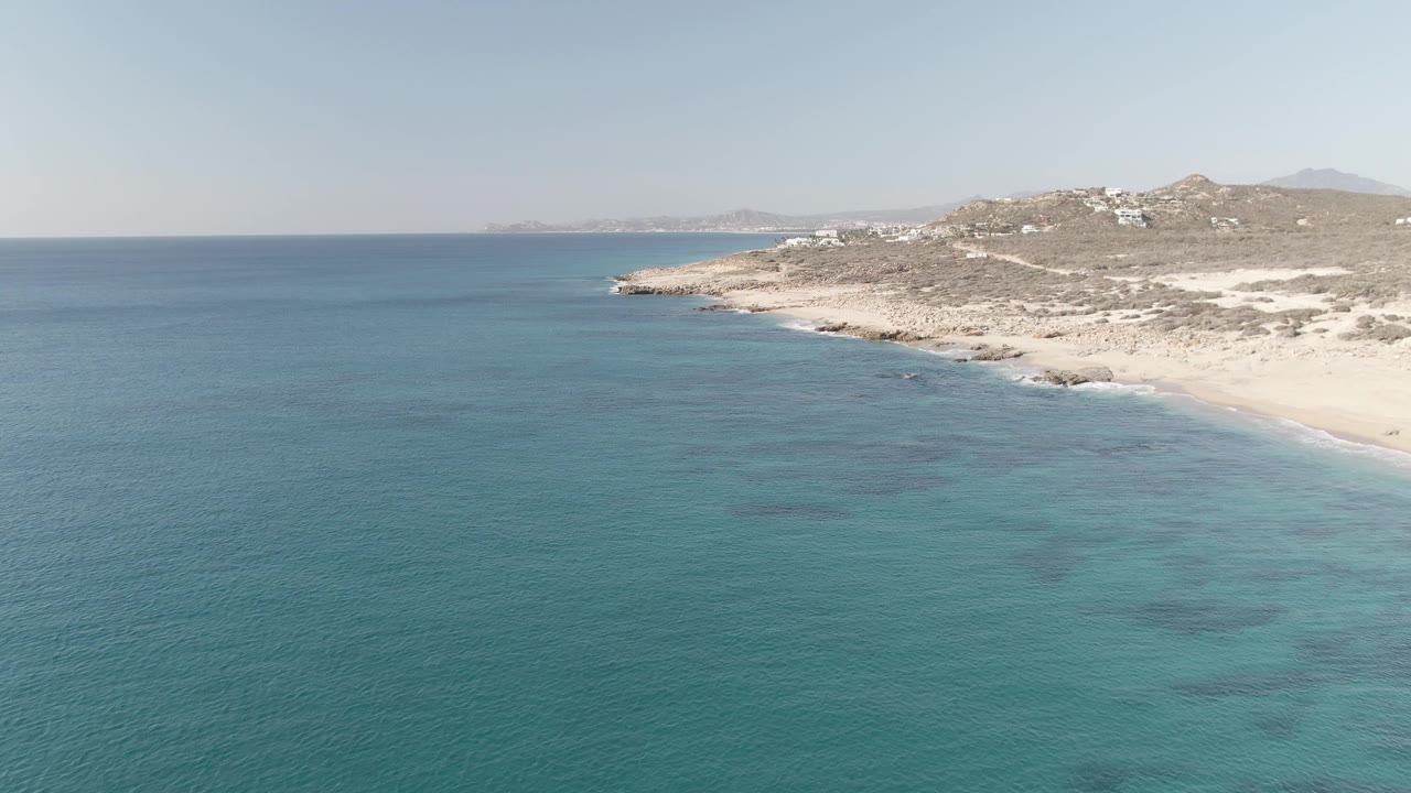 Wide aerial view of blue-green ocean and pale beach below rocky hills.