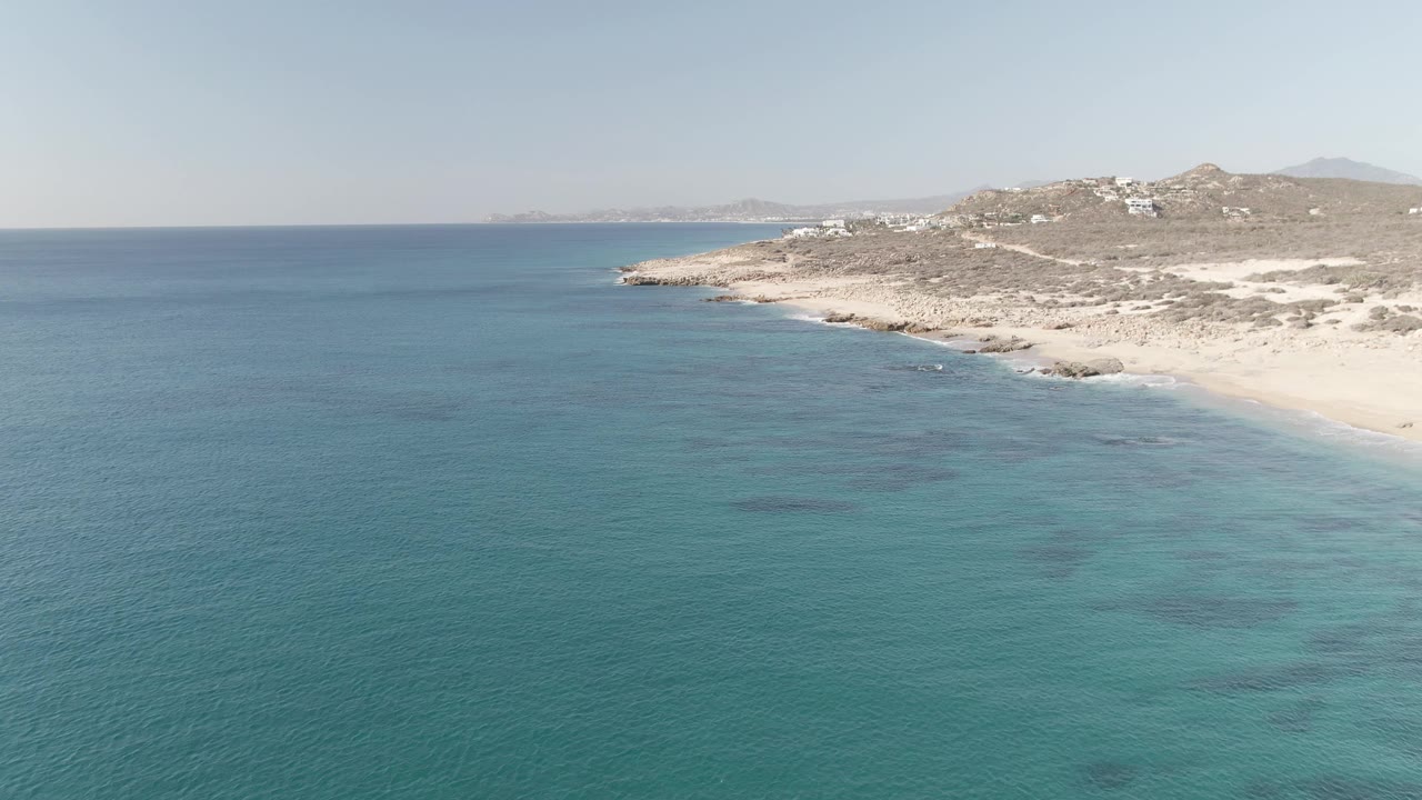 Aerial frame at 0:02 over Buzzards Beach East Cape BCS Mexico showing turquoise shallows closer to shore, small wave lines along the sand, rocky hills, white buildings, and no visible people.