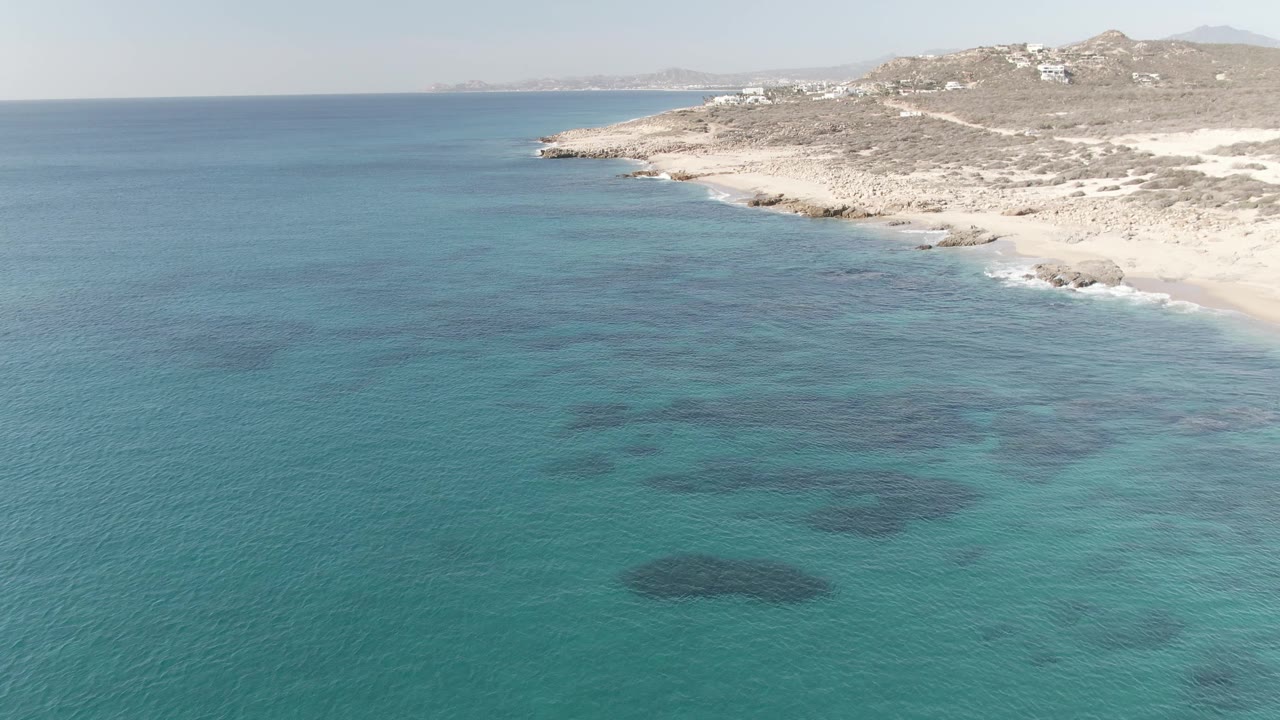 Aerial view at Buzzards Beach East Cape BCS Mexico showing turquoise shallows, dark underwater patches, a pale rocky beach, hillside buildings, and no visible people.