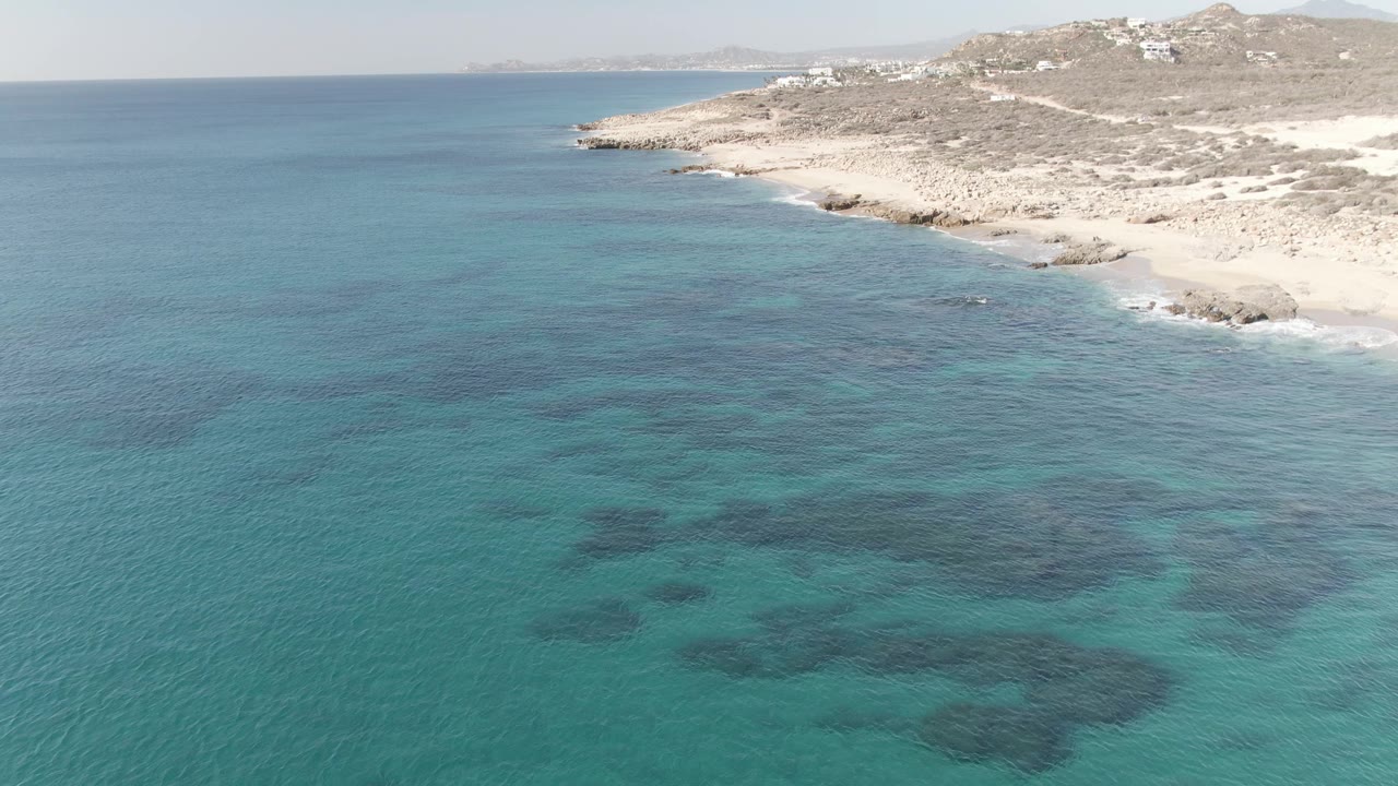 Wide aerial view of Buzzards Beach East Cape BCS Mexico showing open turquoise water in the foreground, a curving rocky shoreline to the right, hillside structures beyond the beach, and no visible people.