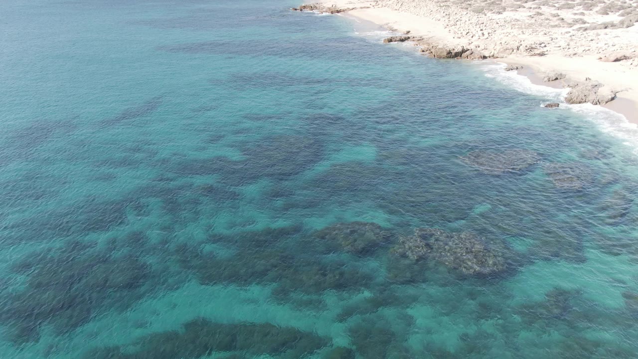From above Buzzards Beach East Cape BCS Mexico, patterned turquoise water fills most of the frame while rough pale rocks and sand trace the upper shoreline, with no visible people.