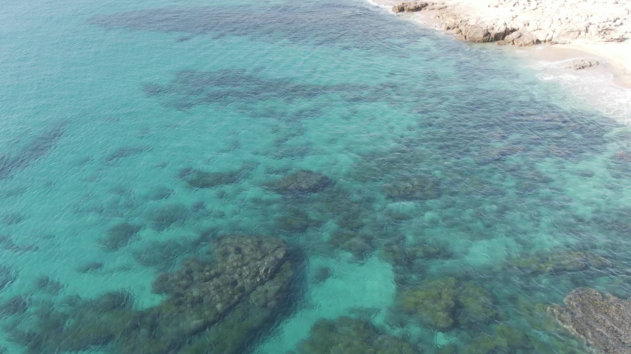 Aerial view at Buzzards Beach East Cape BCS Mexico showing turquoise shallow water over dark underwater rocks, with pale rocky shore at the top right and no visible people.
