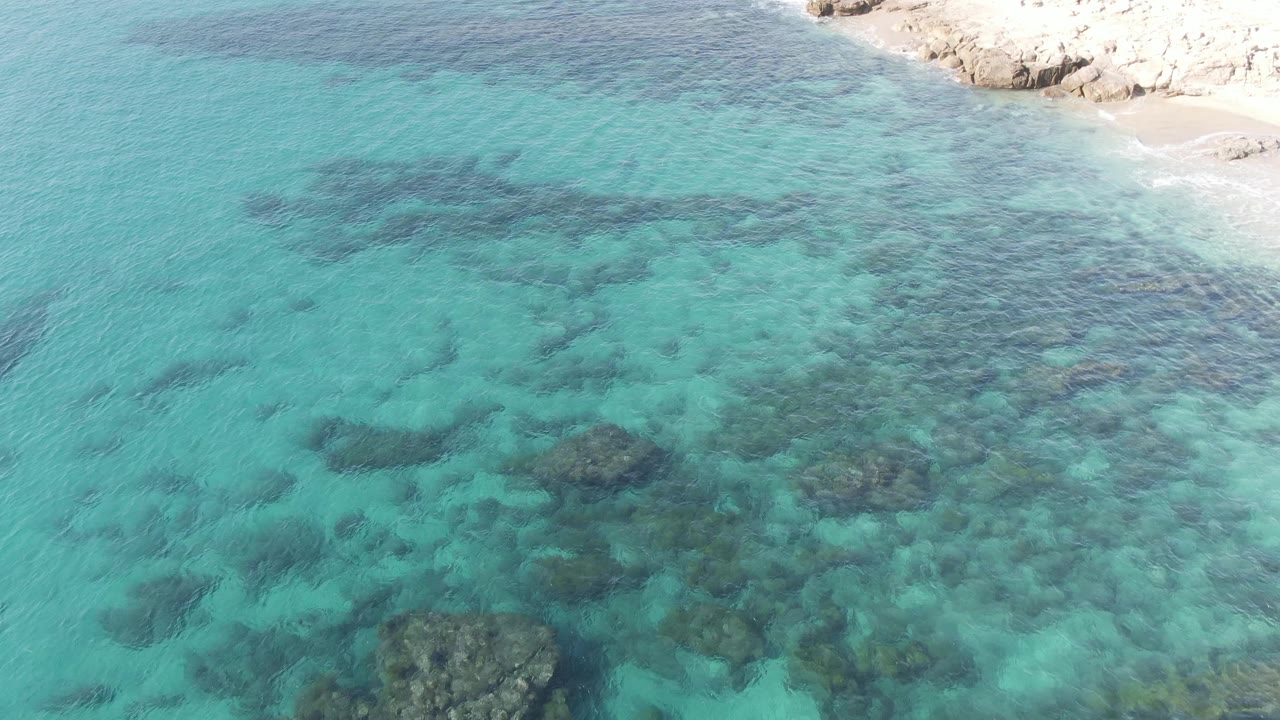Aerial view at Buzzards Beach East Cape BCS Mexico with clear blue-green water, scattered dark rock patches below the surface, a rocky shore along the upper edge, and no visible people.