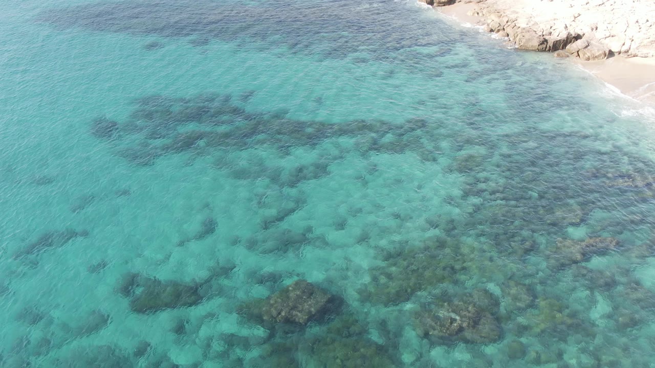 Aerial frame at Buzzards Beach East Cape BCS Mexico showing a wider spread of turquoise water, larger dark submerged rocks across the middle, pale shoreline at the top, and no visible people.