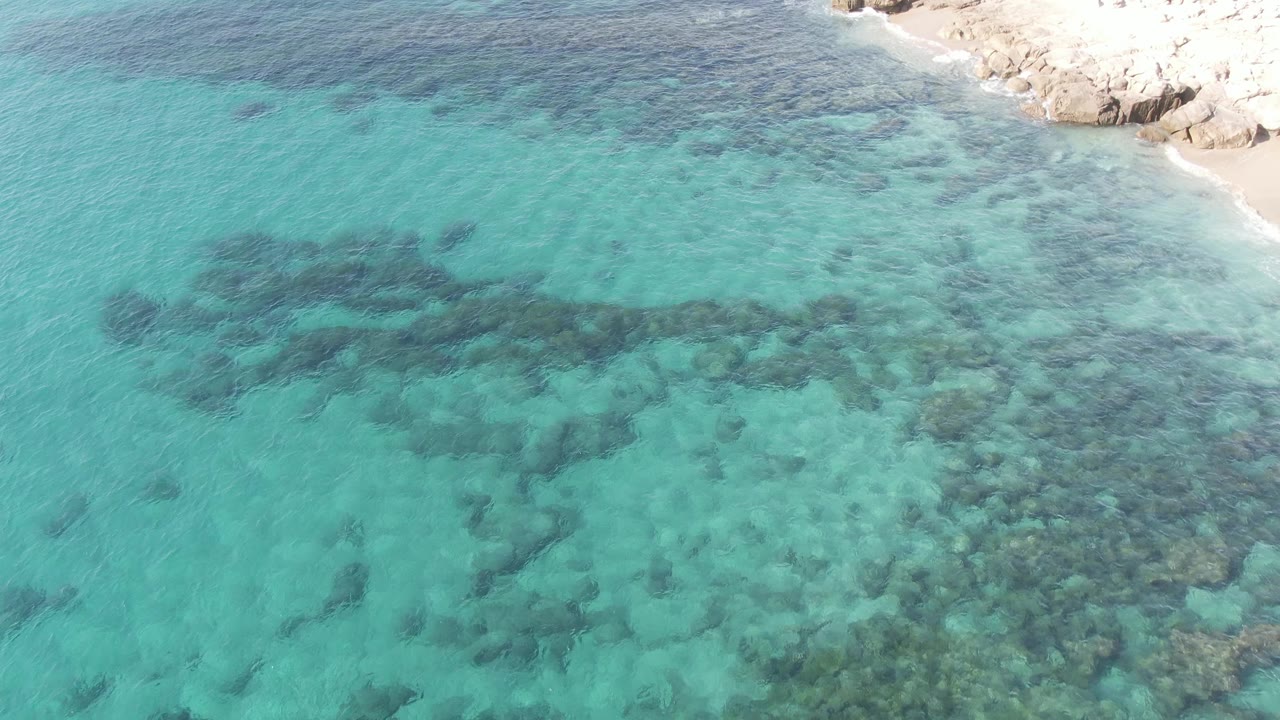 Shallow turquoise ocean with dark underwater rocks and shoreline near the upper right.