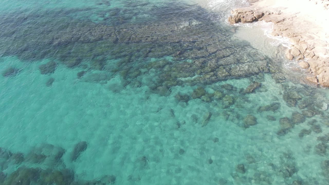 Turquoise shallow water over a dark rocky seabed beside pale sand and shoreline rocks.