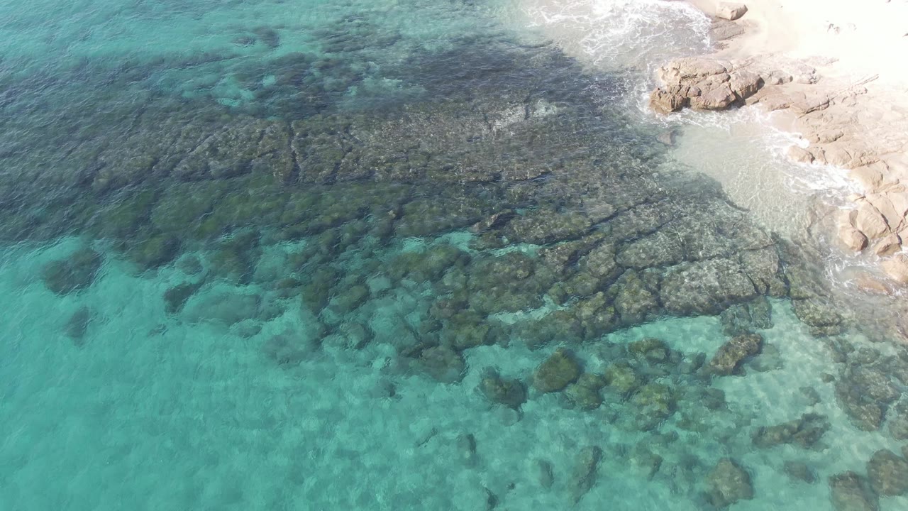 Aerial frame from Buzzards Beach East Cape BCS Mexico with broad submerged rock slabs, turquoise shallows, a narrow sandy shoreline, tan rocks, and no visible people.
