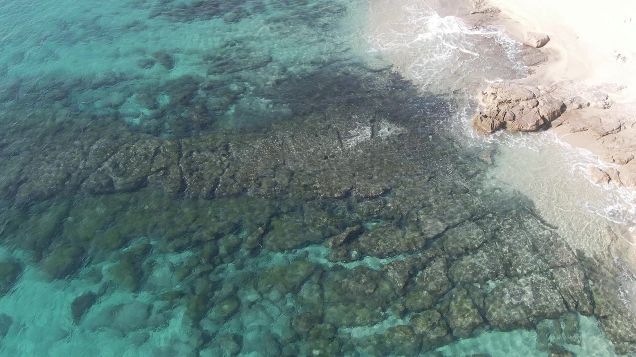Aerial view over Buzzards Beach East Cape BCS Mexico where dark reef-like rocks fill the shallows, small white waves meet pale sand, and no visible people appear.