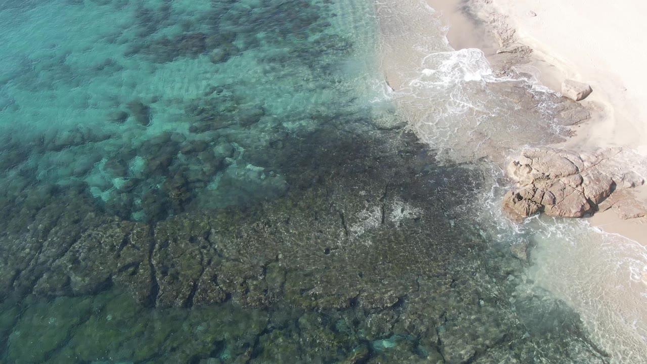 Aerial frame at Buzzards Beach East Cape BCS Mexico showing a darker submerged rock field, clear green-blue water, foamy shore wash on pale sand, and no visible people.