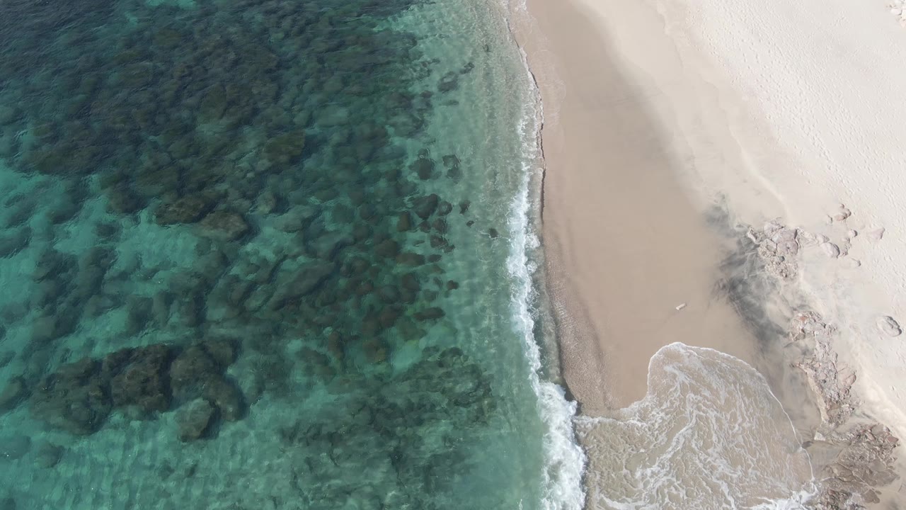 Top-down aerial frame at Buzzards Beach East Cape BCS Mexico showing turquoise shallows, dark submerged rocks, pale sand, a small wash of surf, and no visible people.