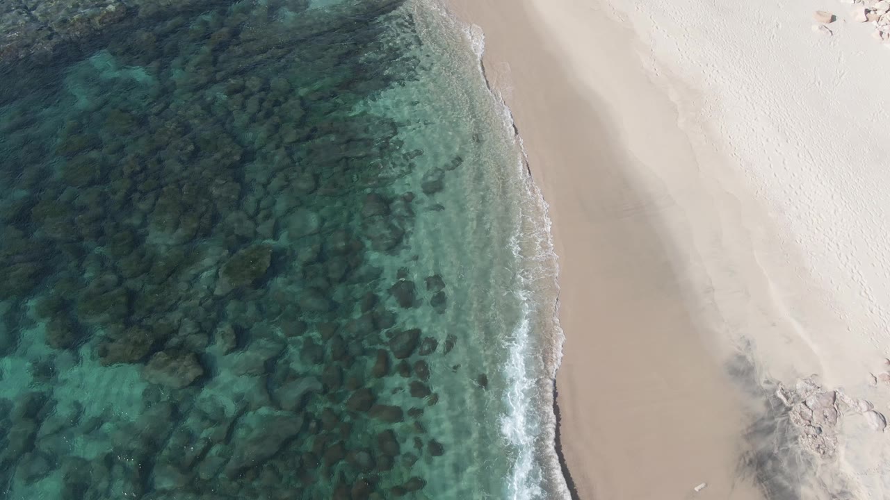 Aerial frame at Buzzards Beach East Cape BCS Mexico with transparent green-blue water over dark rocks, a curved wet shoreline, pale dry sand, and no visible people.