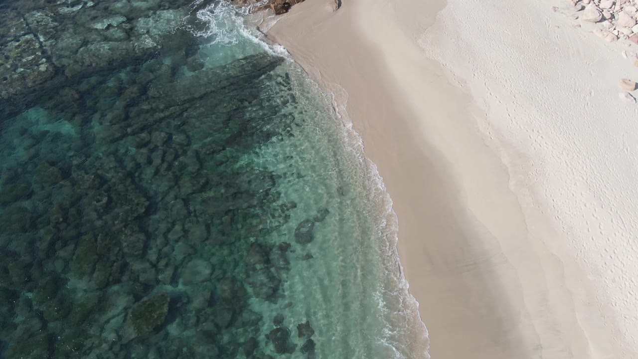 Top-down aerial view at Buzzards Beach East Cape BCS Mexico showing darker rocky water, a bright sand strip, small foamy wave edges, and no visible people.