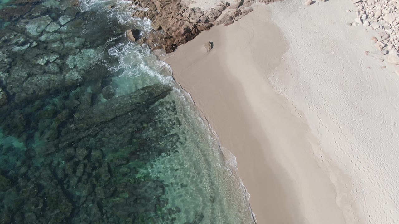 Aerial frame at Buzzards Beach East Cape BCS Mexico showing the beach angled through frame, dark rock patterns below clear water, pale sand, and no visible people.