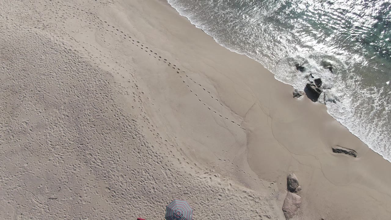 Overhead view of Buzzards Beach East Cape BCS Mexico with pale sand, diagonal footprints, bright surf foam around dark rocks, and a partial striped umbrella at the lower edge, with no visible people.