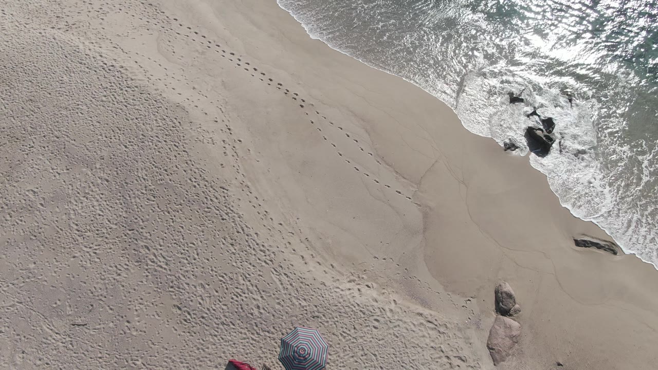Aerial frame of Buzzards Beach East Cape BCS Mexico where the surf foam has shifted around the rocks and the diagonal footprint trail remains clear across the sand, with no visible people.
