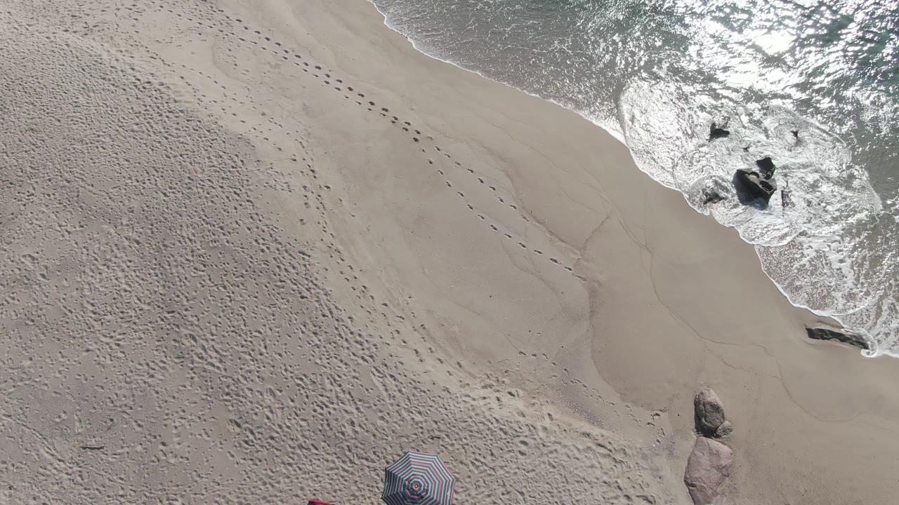 Top-down view at Buzzards Beach East Cape BCS Mexico with bright reflected water on the upper right, foam surrounding rocks, textured sand, and the striped umbrella partly visible at the bottom, with no visible people.