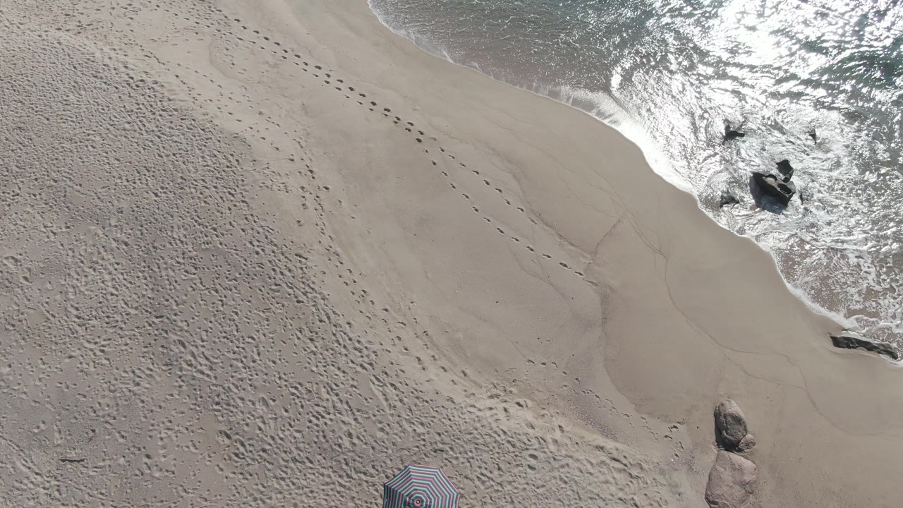Overhead beach sand with footprints, foamy shore break, rocks, and partial umbrella.