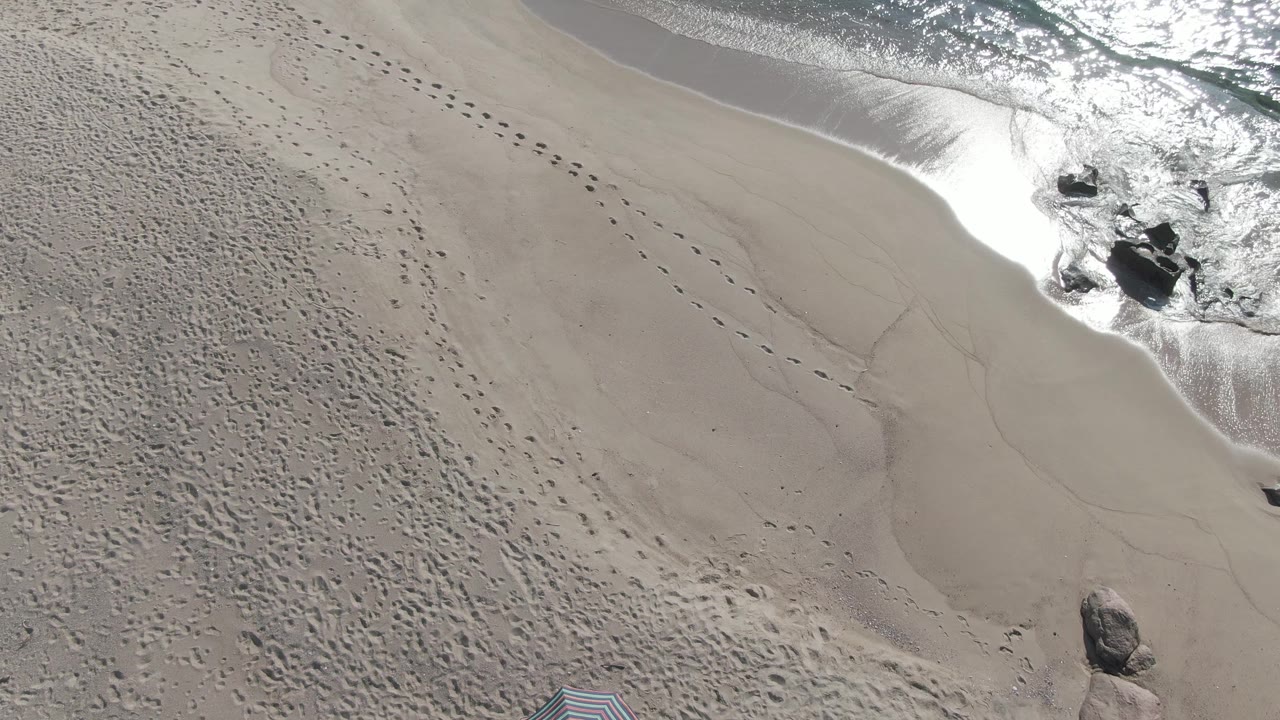 Overhead view of Buzzards Beach East Cape BCS Mexico showing pale sand, scattered footprints, dark shoreline rocks, white surf foam, and no visible people.
