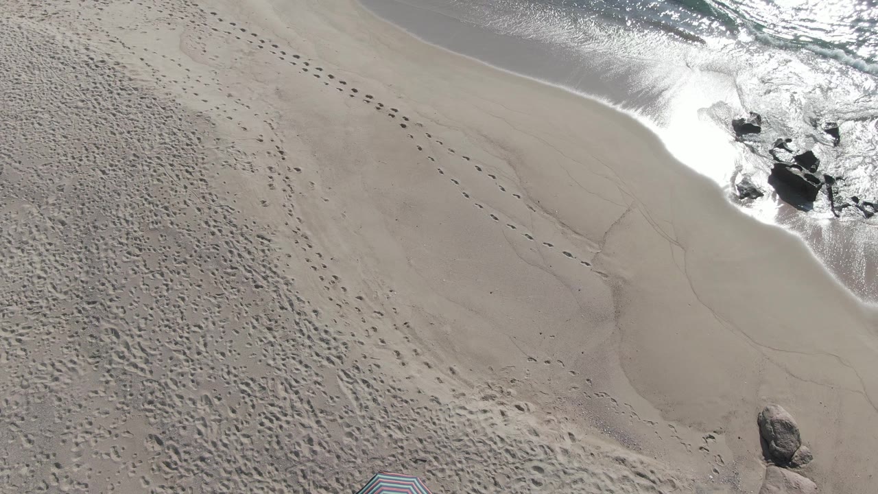 Aerial frame from Buzzards Beach East Cape BCS Mexico with footprint trails across dry sand and waves wrapping around dark rocks near the top of frame, with no visible people.