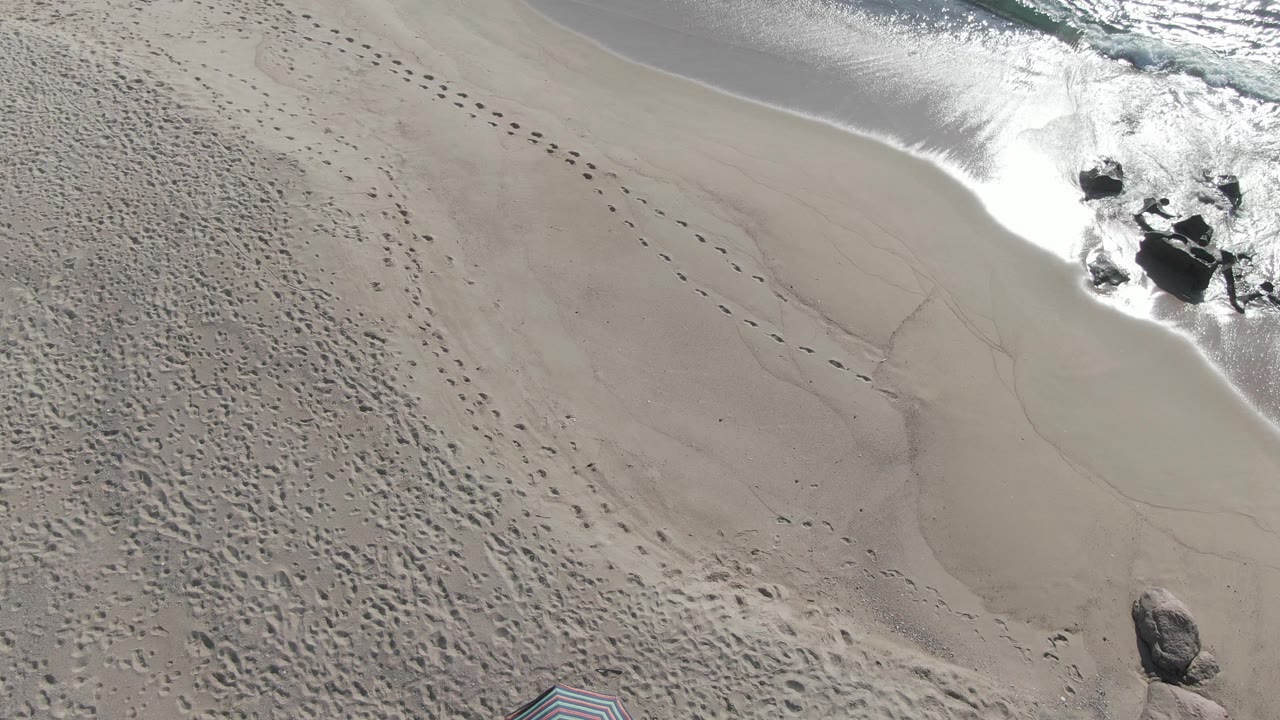 Overhead Buzzards Beach East Cape BCS Mexico view with a diagonal surf edge, clustered dark rocks, pale sand marked by footprints, and no visible people.