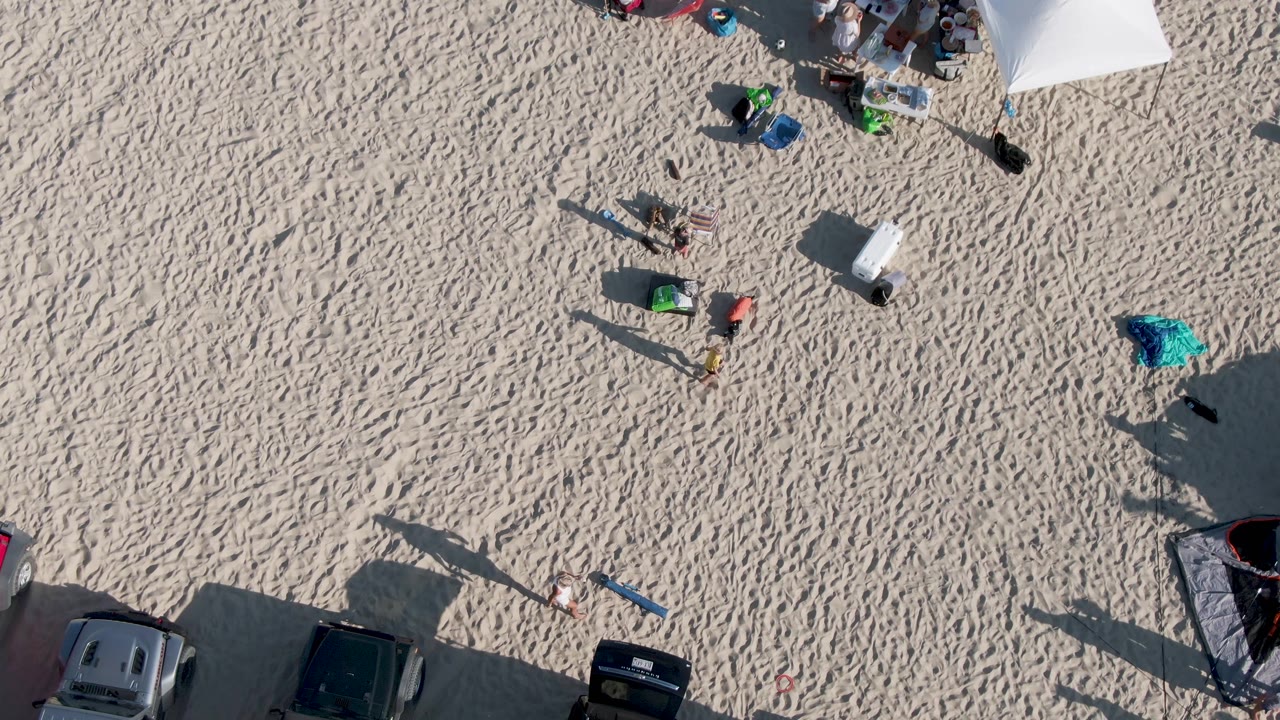 Overhead frame at Shipwrecks Beach in Mexico showing rippled sand, several people near beach gear, the edge of a white tent, and parked cars along the bottom.