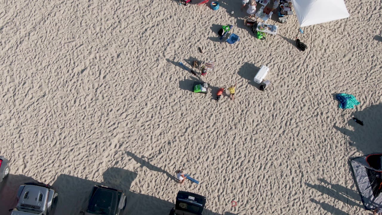 Aerial beach view with small figures, chairs, coolers, tent corner, and cars.