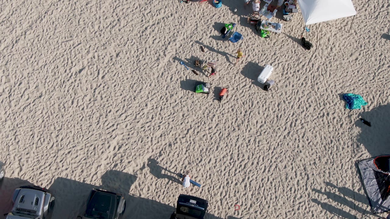 Top-down frame at Shipwrecks Beach in Mexico showing a cluster of people and items near the upper edge, open rippled sand across the center, and parked vehicles along the lower edge.
