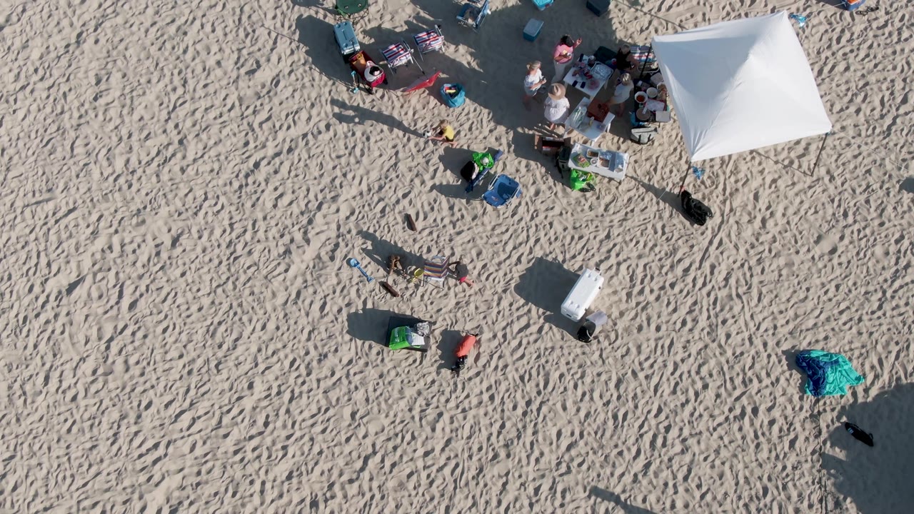 Overhead view at Shipwrecks Beach in Mexico showing a white shade tent, beachgoers, chairs, coolers, towels, and long shadows on rippled sand.