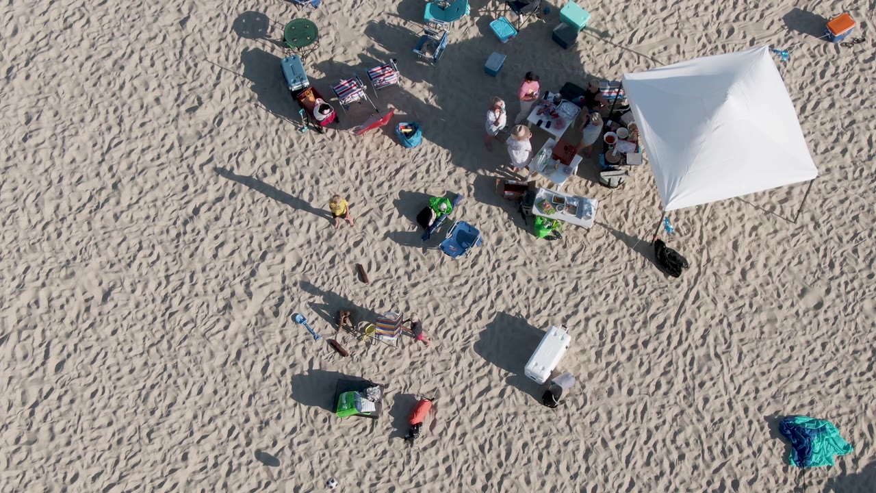 Top-down Shipwrecks Beach frame showing the white tent on the right, people gathered at its edge, and loose beach gear spread across the sand.
