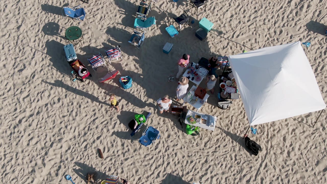 Overhead frame from Shipwrecks Beach in Mexico showing a white canopy at right, scattered folding chairs and coolers on footprint-marked sand, and several visible beachgoers around tables.