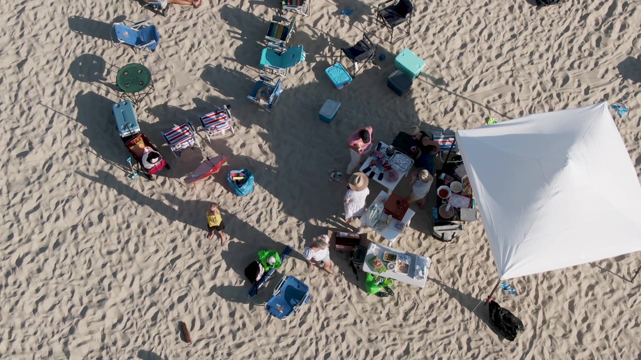 Beachgoers stand and move near tables beside a white tent on sandy beach.