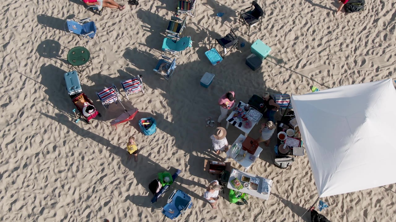 At 0:02, the Shipwrecks Beach frame keeps the white tent at right while chairs, coolers, bags, and beachgoers form a loose campsite pattern across the sand.