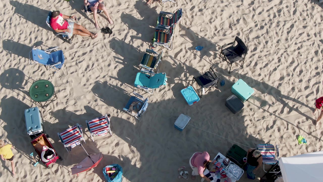 Overhead frame at Shipwrecks Beach in Mexico showing beachgoers seated and reclining among folding chairs, coolers, bags, and long shadows on pale sand.