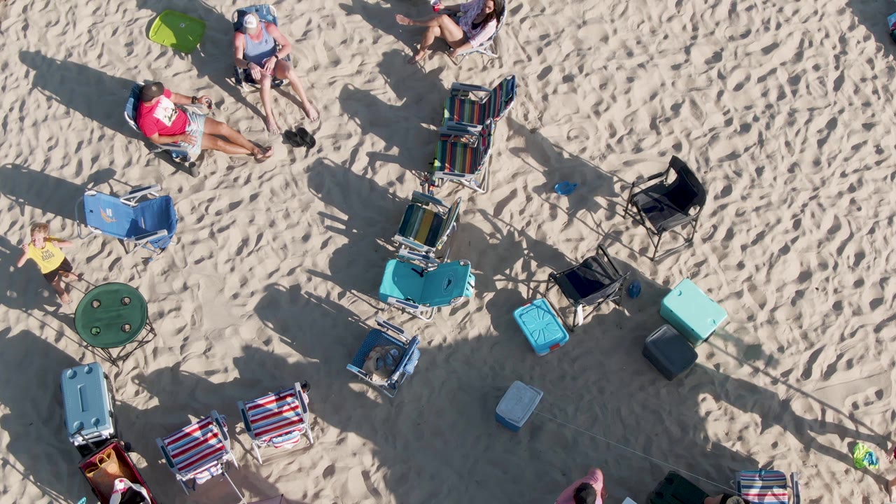 At 0:02, the aerial view over Shipwrecks Beach in Mexico shows adults relaxing on chairs and towels, scattered coolers and bags, a green umbrella edge, and dark shadows on rippled sand.