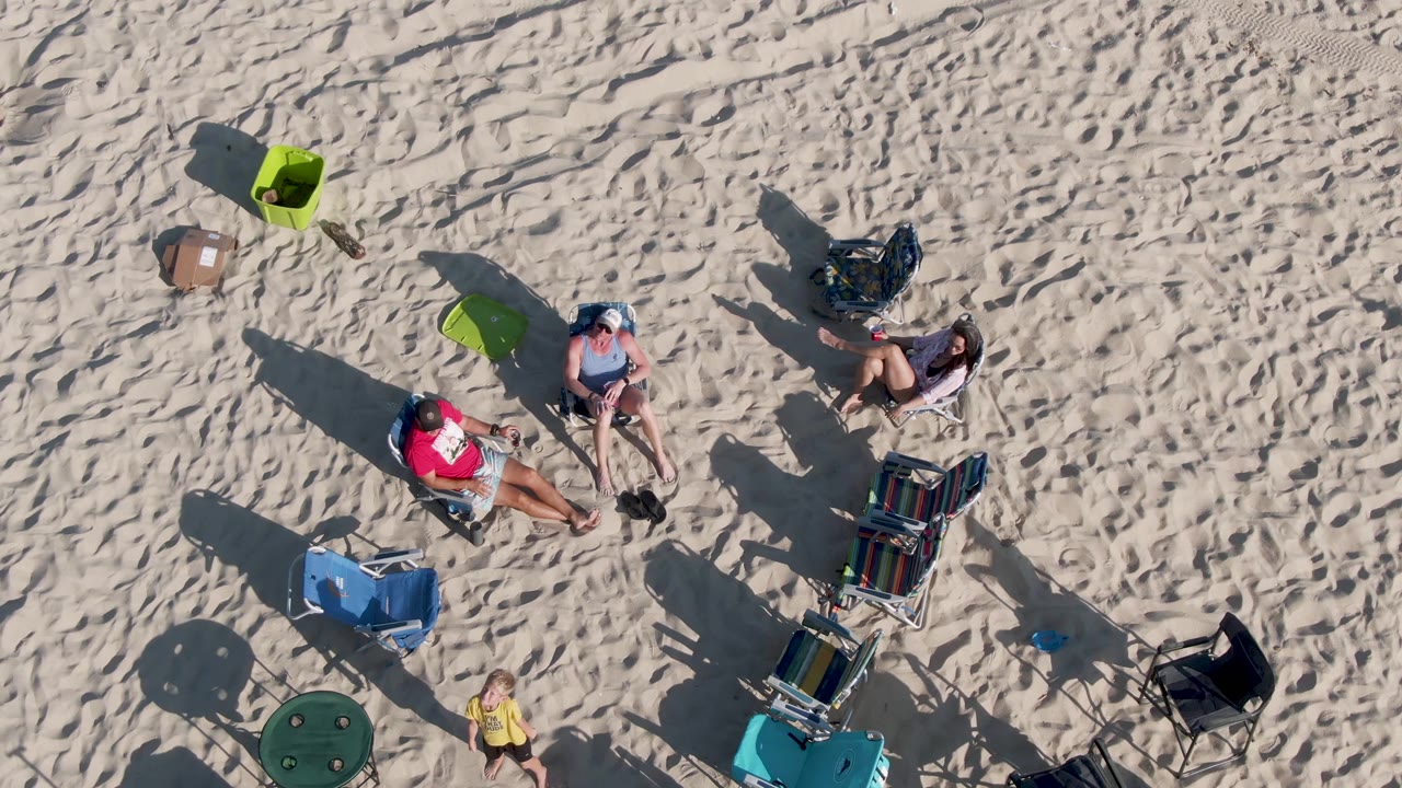 Overhead frame from Shipwrecks Beach East Cape Los Cabos BCS Mexico showing beachgoers on pale sand, a child near the lower edge, folding chairs, towels, and long shadows.