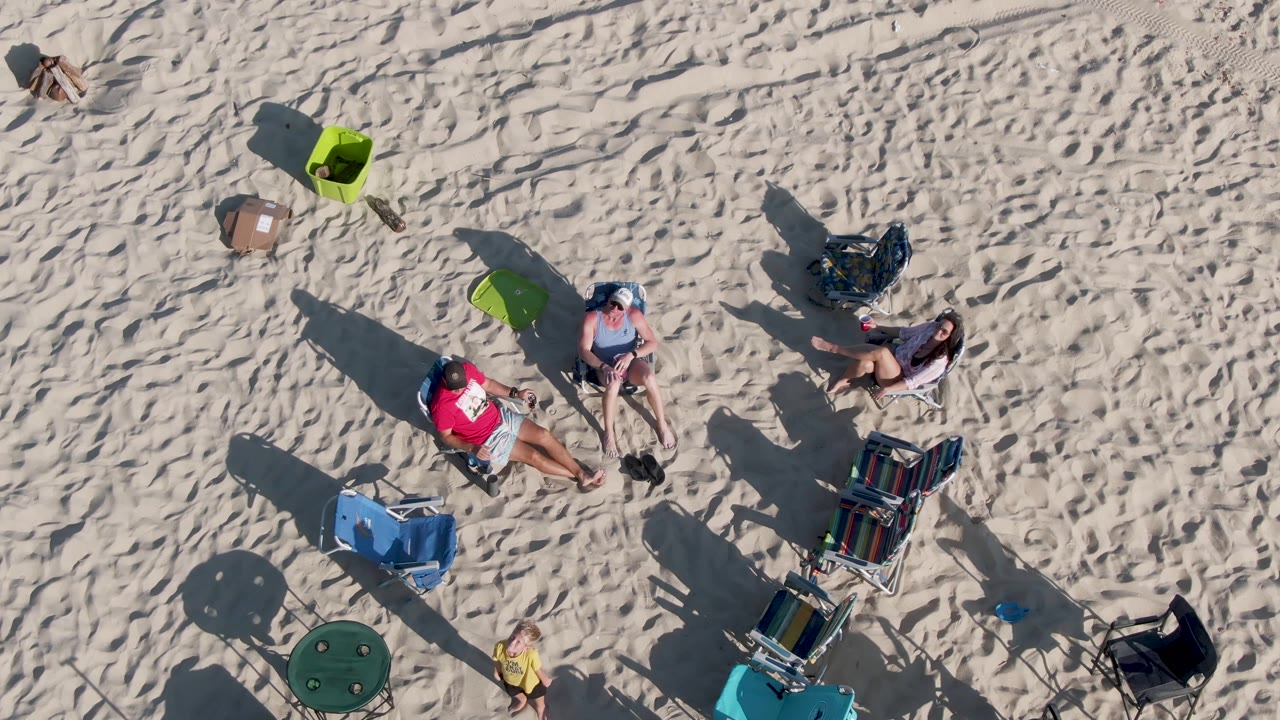 Beachgoers sit and recline on sand beside folding chairs and coolers.