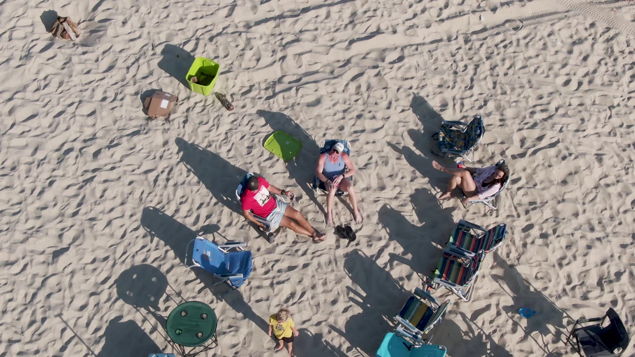 At 0:02, people remain seated on the sandy beach in Mexico while the yellow-shirted child appears near the lower edge among chairs, towels, coolers, and dark angled shadows.