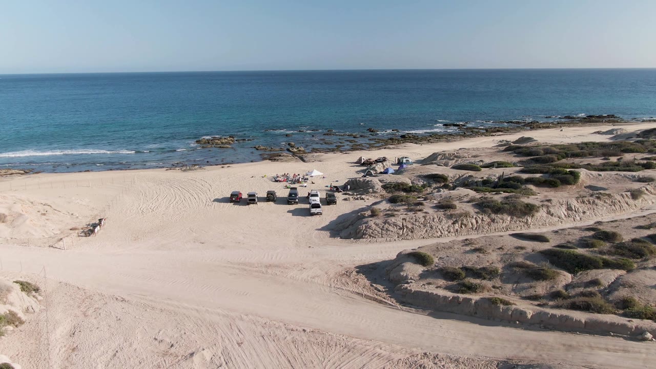 Third aerial frame over Shipwrecks Beach East Cape Los Cabos with the vehicles and umbrellas still visible on open sand, wave lines reaching the rocks, and no visible people.
