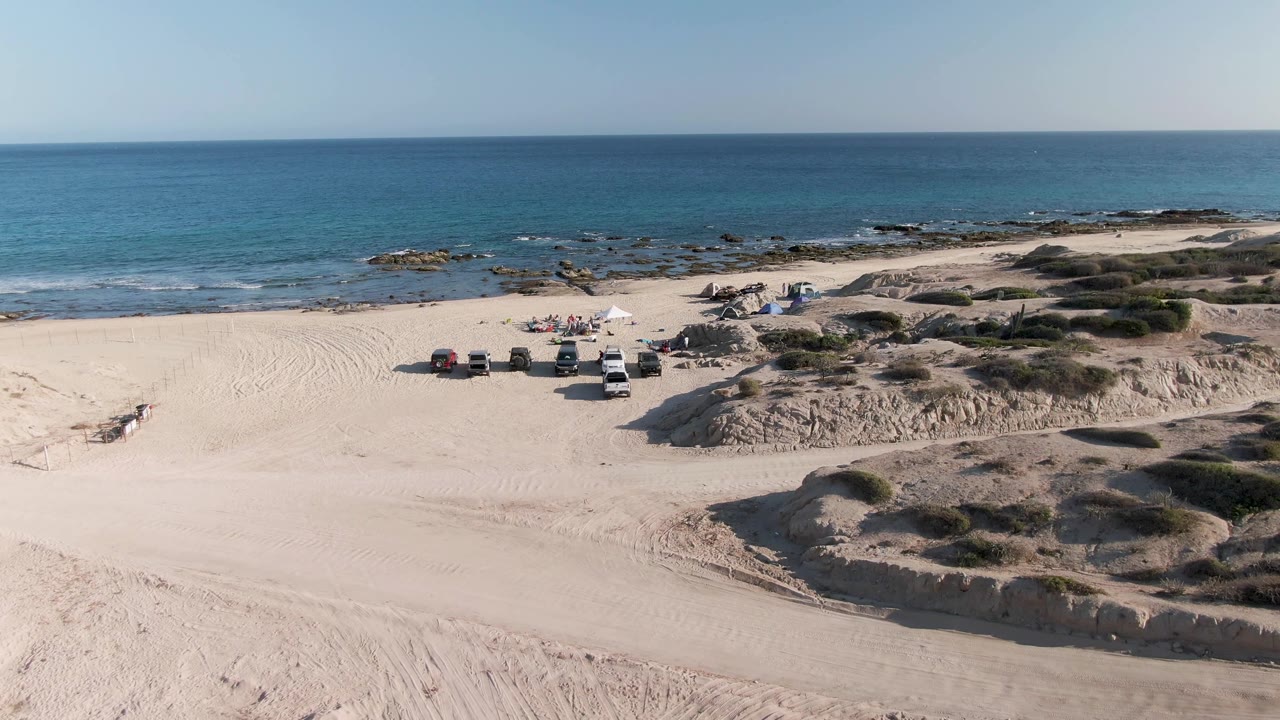 Aerial frame at Shipwrecks Beach East Cape Los Cabos BCS Mexico showing pale sand dunes, several vehicles near a small campsite, distant people, rocky shoreline, and blue ocean.