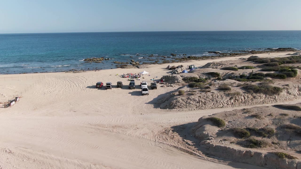 Aerial frame at Shipwrecks Beach East Cape Los Cabos BCS Mexico showing the campsite and vehicles slightly farther right in the composition, with pale dunes in front and blue water beyond.