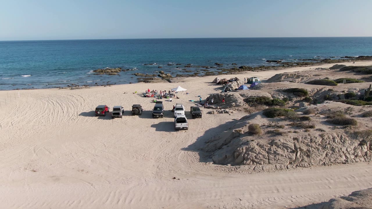 Aerial frame at Shipwrecks Beach shows several vehicles lined up on pale sand, people gathered near a white canopy, rocky shore, and blue ocean beyond.