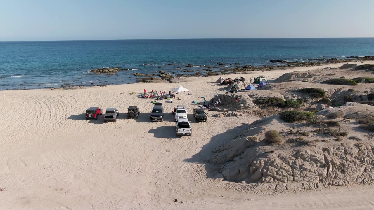 Vehicles sit on a sandy beach with people and tents near the shoreline.